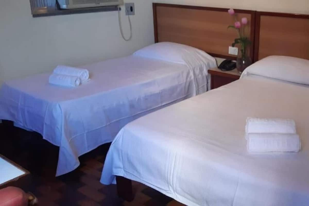 Habitación de hotel con dos camas individuales, cada una con una colcha blanca y una toalla doblada. Se observa un mesita de noche con un teléfono y un florero con flores.