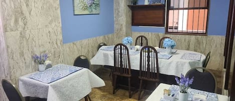 A dining area with tables set for four, each with a white tablecloth and blue patterned placemats. There are chairs around the tables, and a wall-mounted shelf with decorative items.