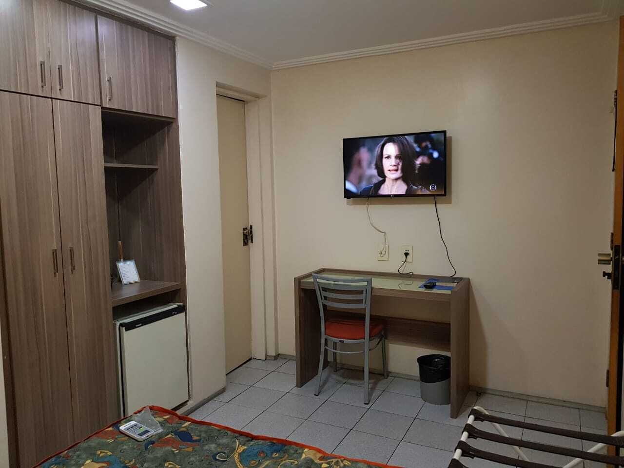 Habitació amb armari de fusta, escriptori amb cadira, televisió penjada a la paret i nevera petita.