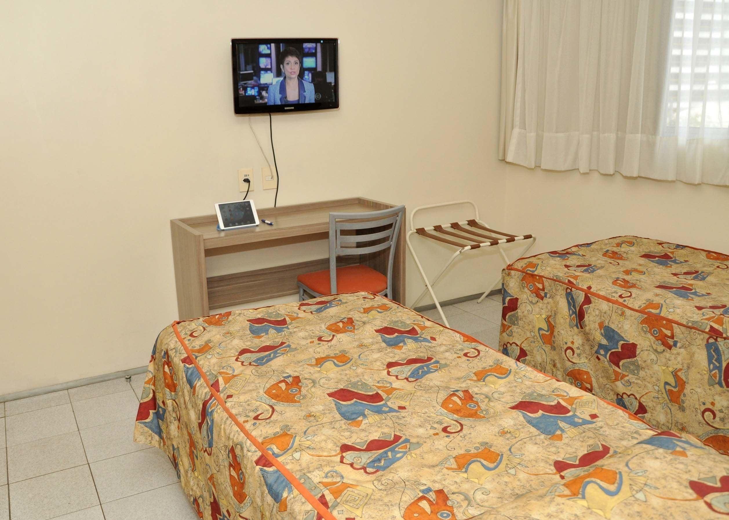 A room with two beds, a desk, a chair, a television, and a window with curtains.