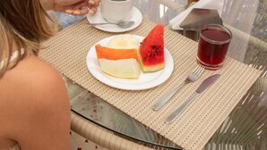 Una persona sentada en una mesa de vidrio con una taza de café, un plato con una tajada de sandía y un vaso de bebida roja.