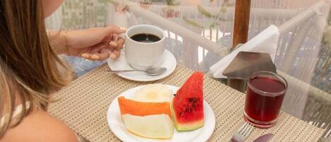 A person seated at a glass table with a cup of coffee, a plate with a slice of watermelon, and a glass of red beverage.