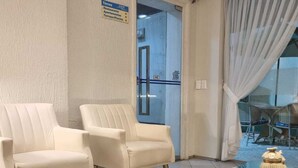 Una sala de estar con dos sillones blancos, una mesa de centro de vidrio y un sofá color beige. Un cartel indica un restaurante, apartamentos y una terraza/piscina.