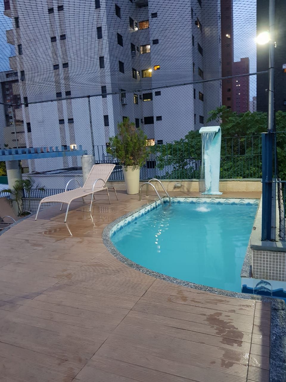 Una piscina exterior con una cascada, rodeada por una valla y sillones de descanso.