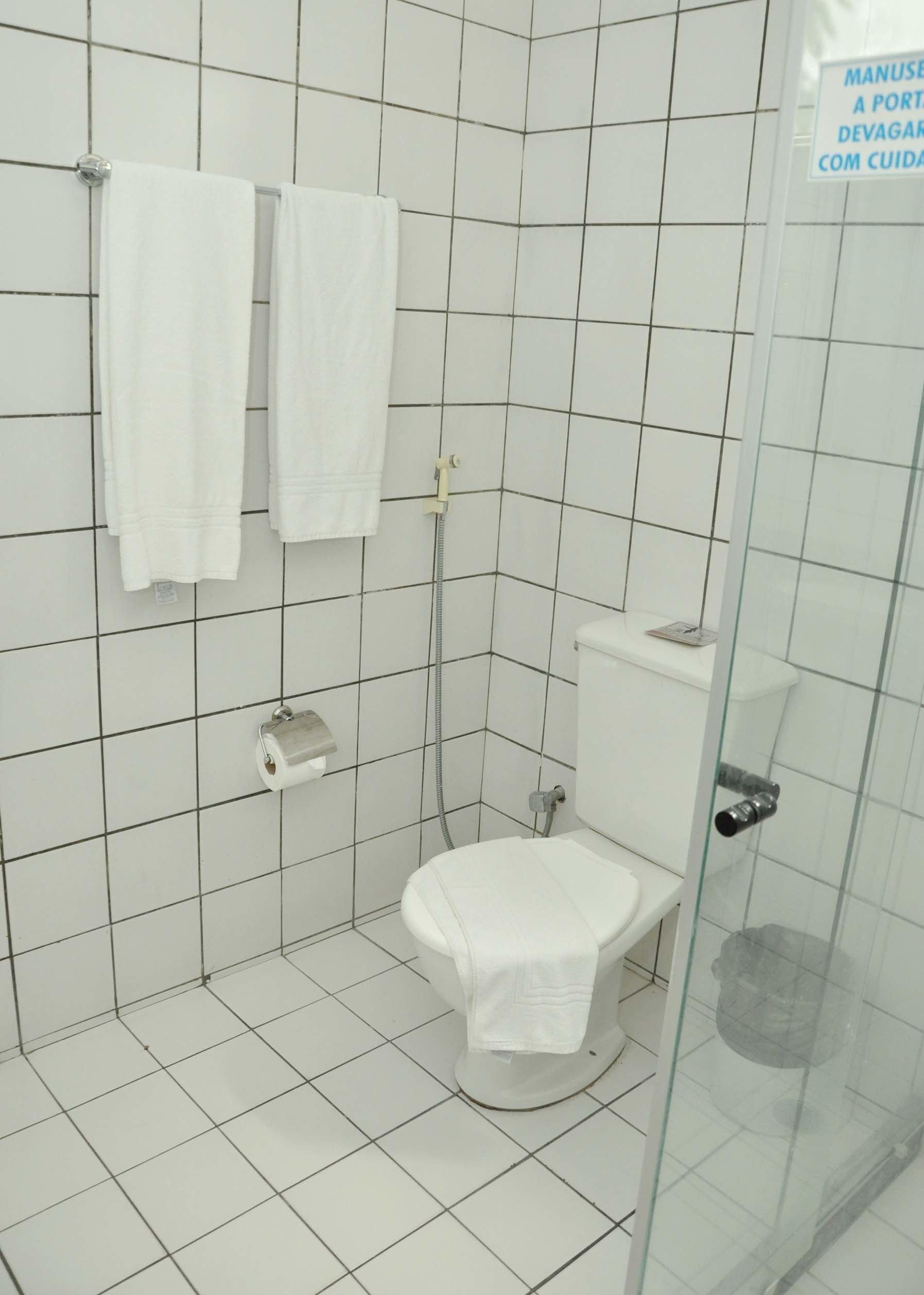 A small, tiled bathroom with a glass shower enclosure, a toilet, and two white towels hanging on the wall.