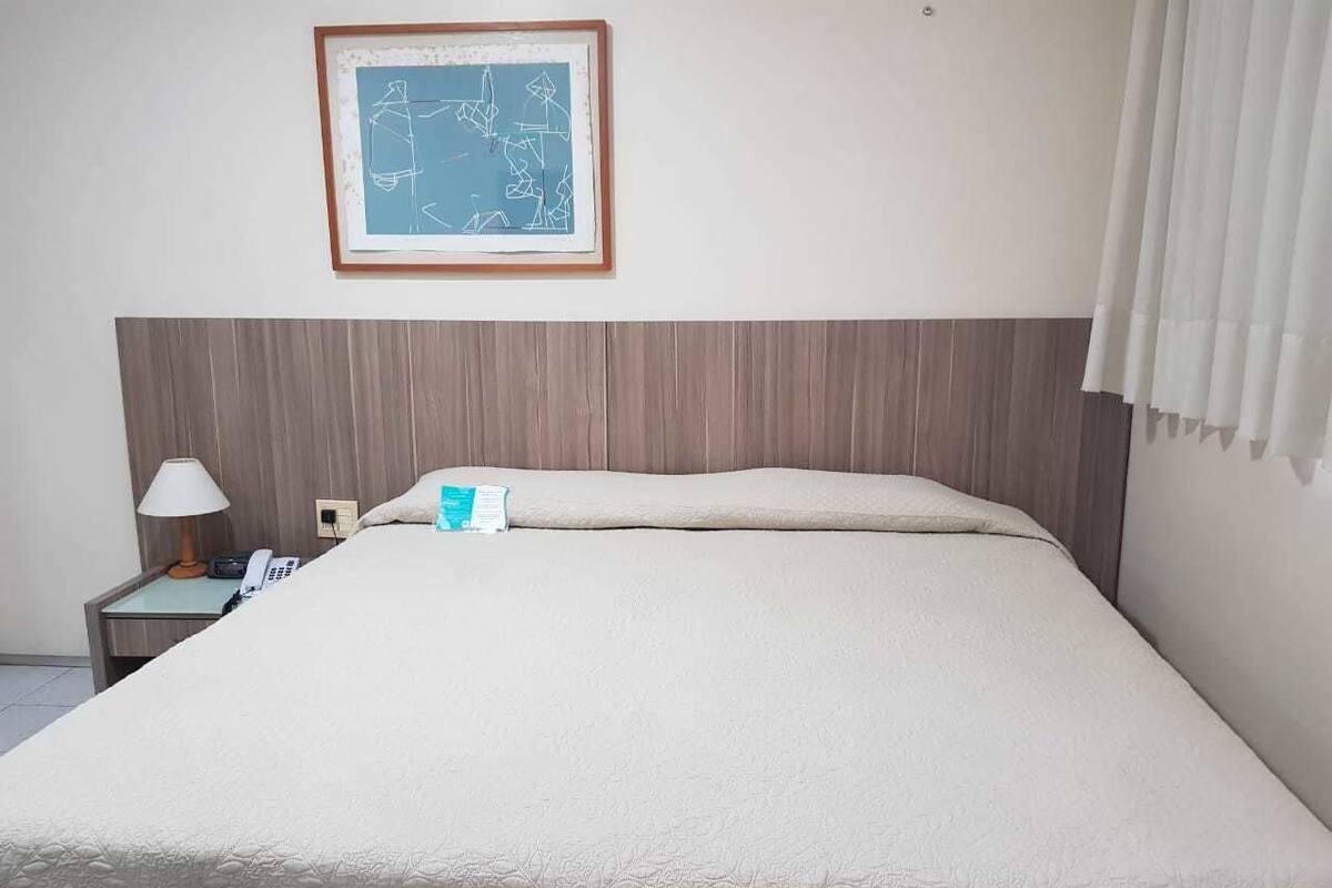 Hotellihuone, jossa on suuri sänky, yöpöytä lampun kanssa, puhelin ja seinällä oleva kehystetty abstrakti taideteos.