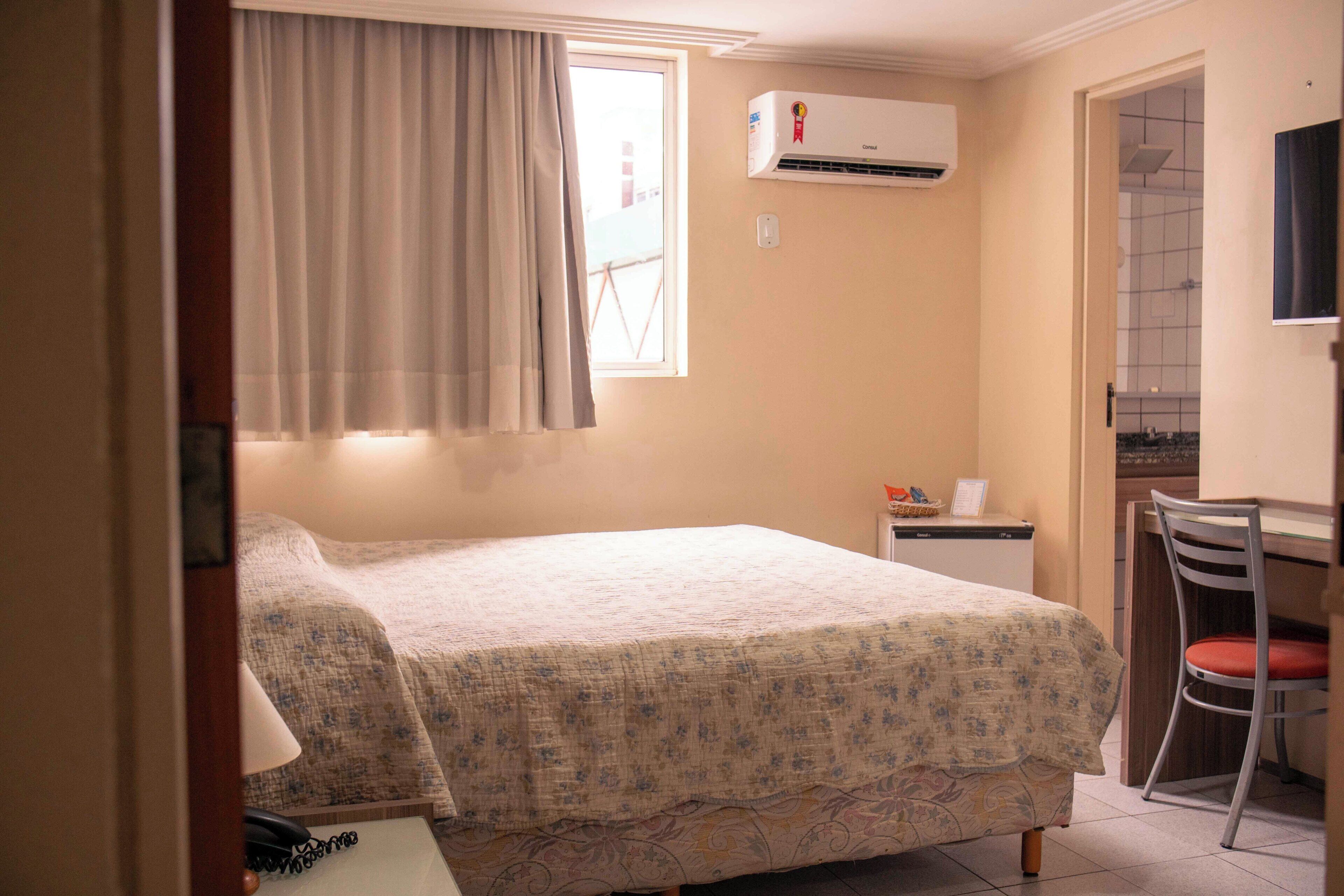 Un dormitorio con una cama, aire acondicionado, un escritorio con silla y una ventana con cortinas.