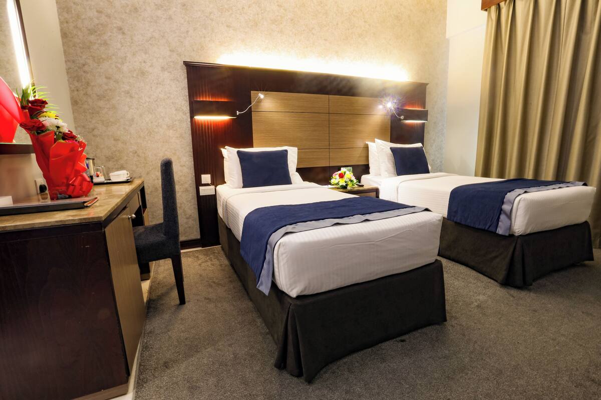 A hotel room with two beds, a desk, a chair, a nightstand, and a lamp.