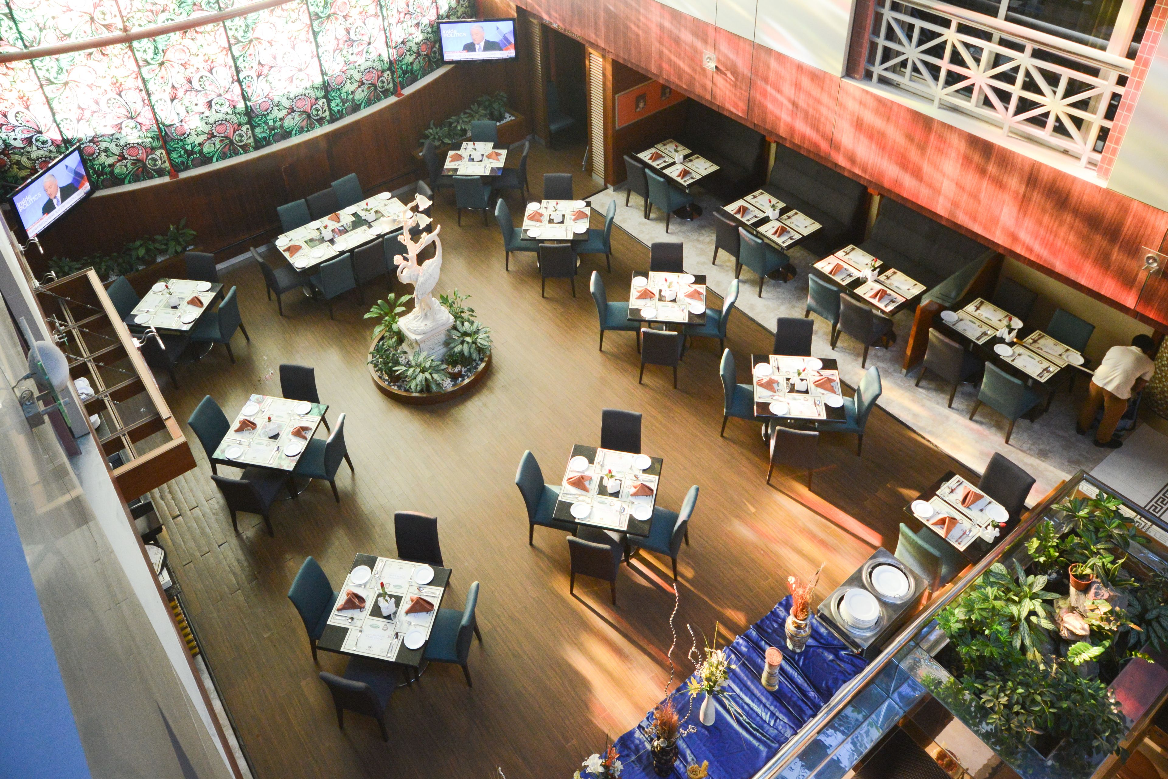 An indoor dining area with multiple tables and chairs, a central decorative plant, and a television screen mounted on the wall.