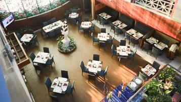 An indoor dining area with multiple tables and chairs, a central decorative plant, and a television screen mounted on the wall.