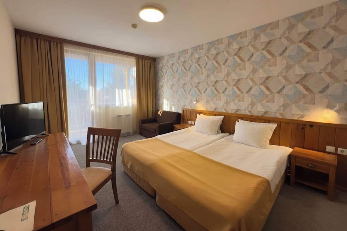 A hotel room with a bed, bedside tables, a desk with a TV, a chair, and a window with curtains.