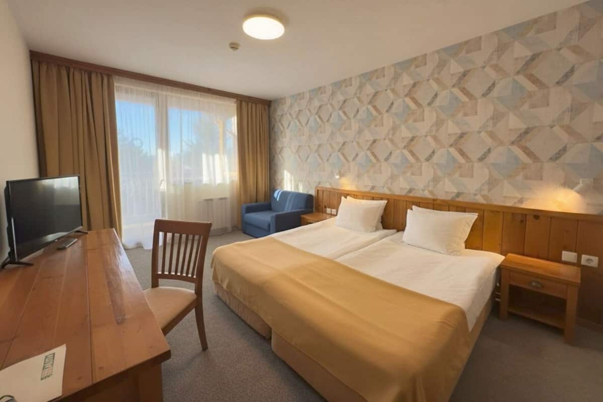 A hotel room with a wooden desk, two beds, a TV, a chair, and a patterned wall.