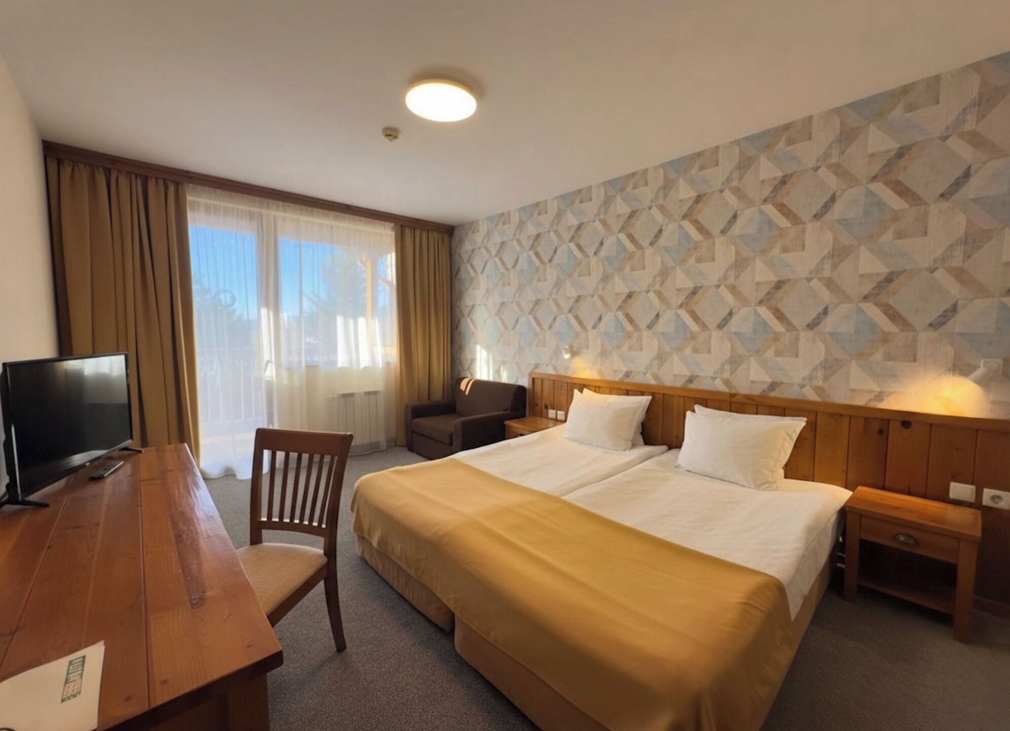 A hotel room with a bed, bedside tables, a desk with a TV, a chair, and a window with curtains.