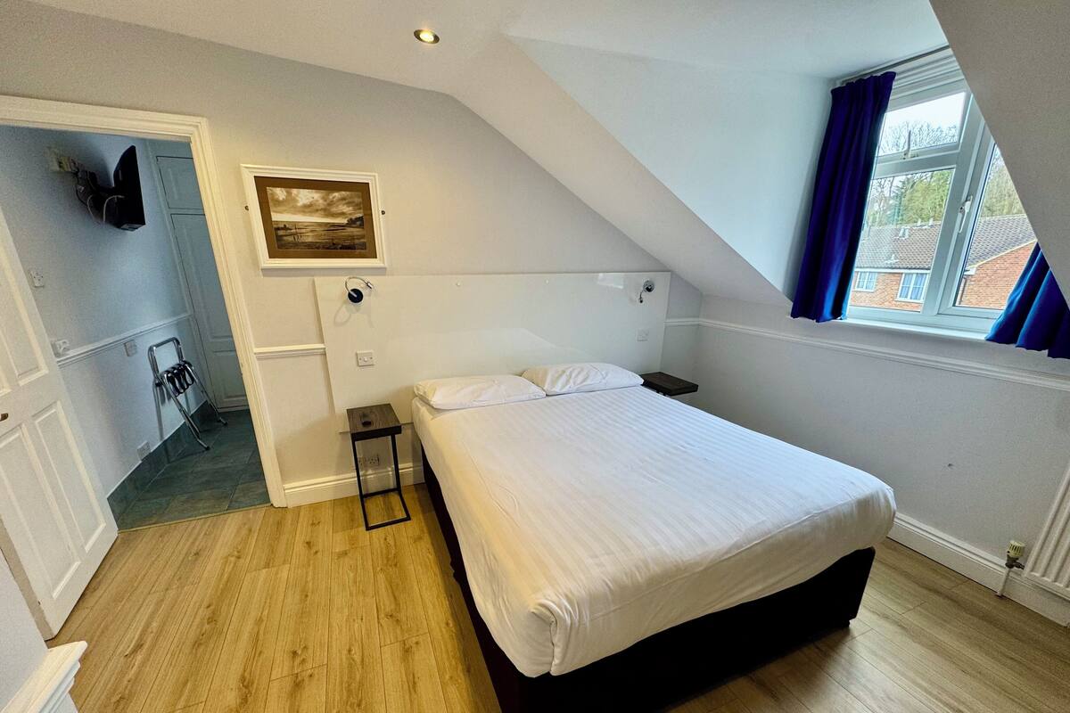 Une chambre à coucher avec un lit, une table de chevet, une petite table, une chaise, un tableau au mur et une fenêtre avec des rideaux.
