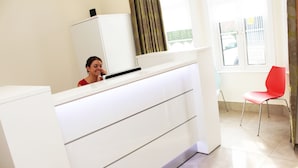 A reception desk with a white counter and drawers, a red chair, and a window with curtains. - Brentwood Hotel (Brentwood)