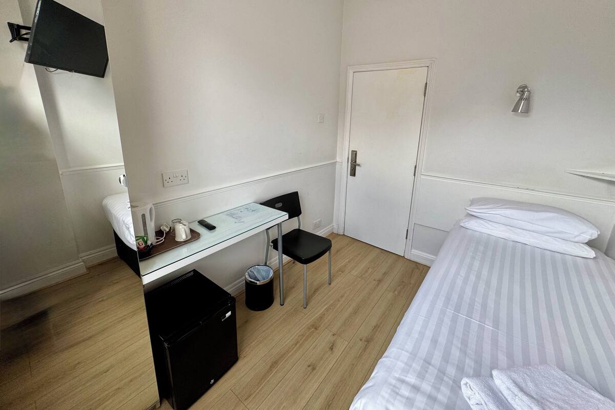 Une chambre d’hôtel de taille réduite, comprenant un lit simple, un petit bureau avec une chaise, une poubelle et une porte blanche.