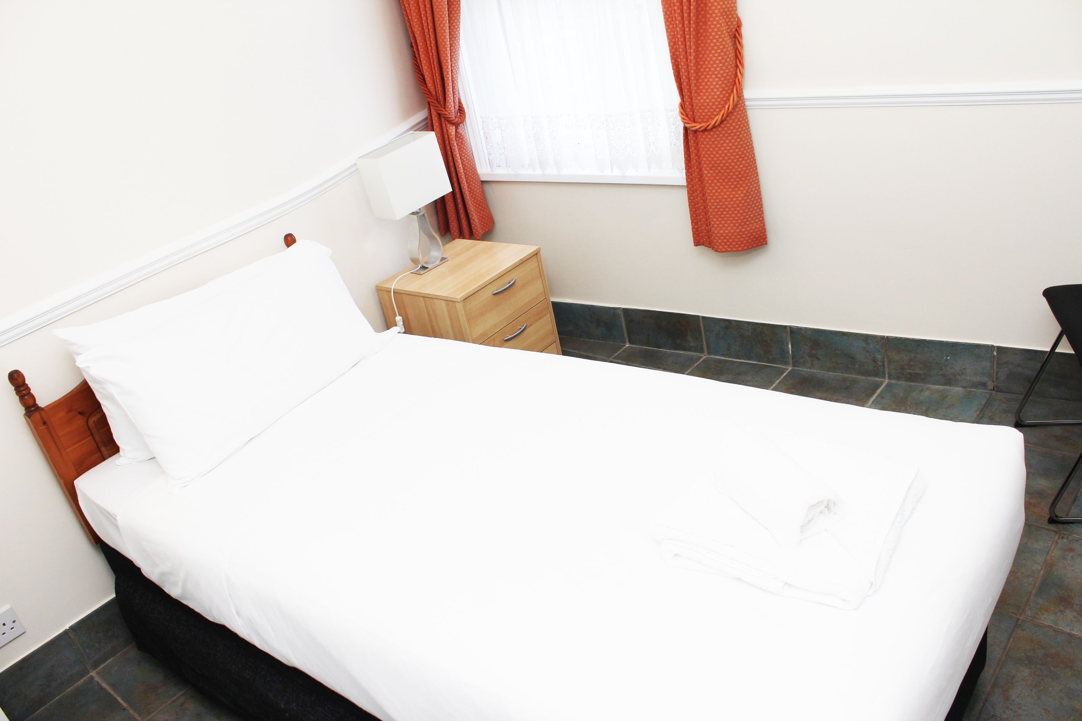 A neatly made bed with white linens, a wooden nightstand with a lamp, and a chair in a room with tiled flooring and a window with orange curtains.