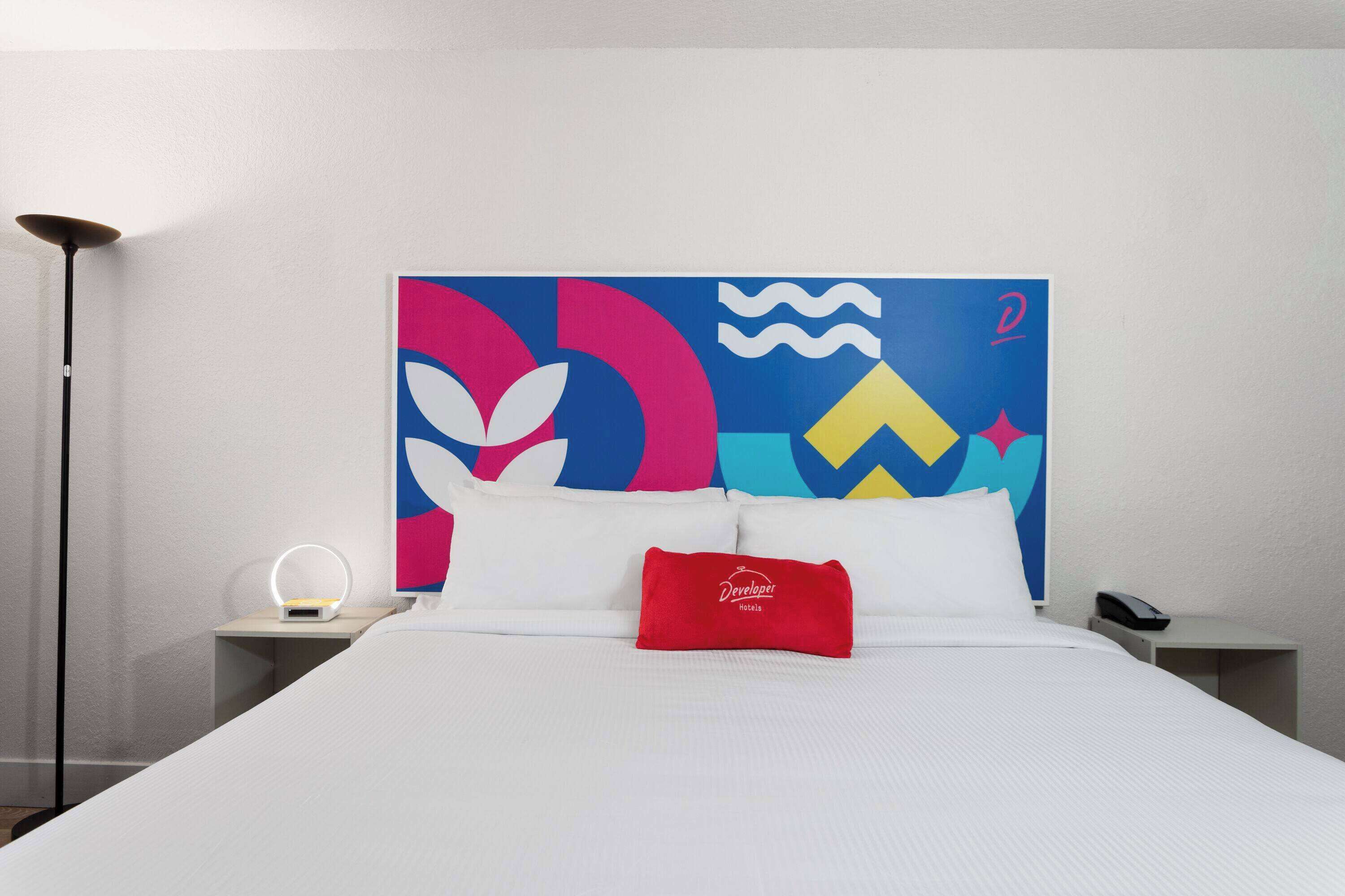 Habitación de hotel con cama, mesita de noche con lámpara, teléfono y un cuadro abstracto de colores en la pared.