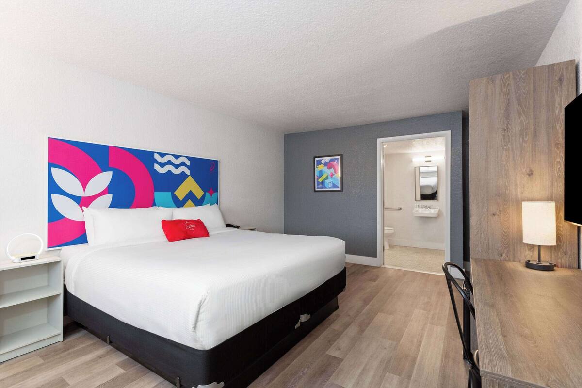 A modern hotel room with a large bed, a bedside table, a desk, and a chair.