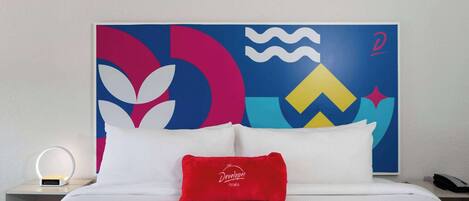 Un cuarto de hotel con cama, ropa de cama blanca, almohada roja, mesita de noche con lámpara y un cuadro abstracto colorido en la pared.