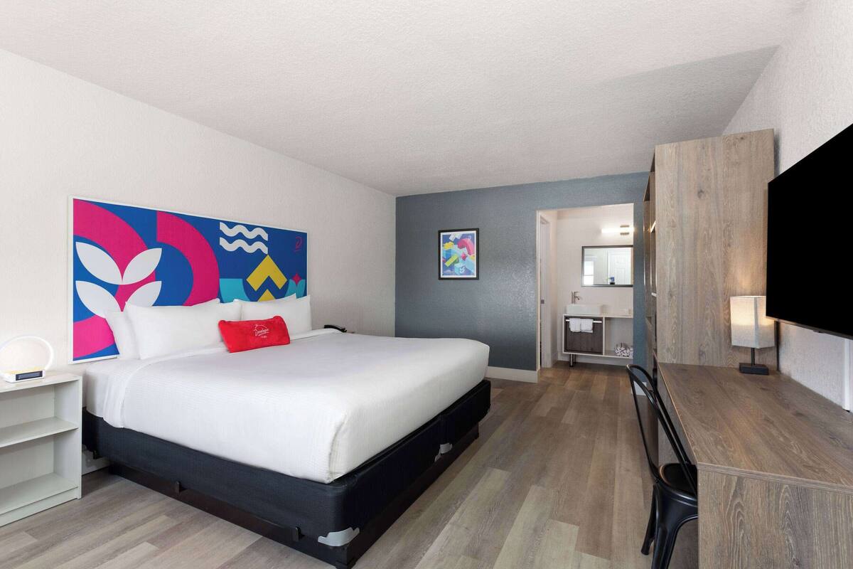 A modern hotel room with a bed, a desk, a TV, and artwork on the walls.