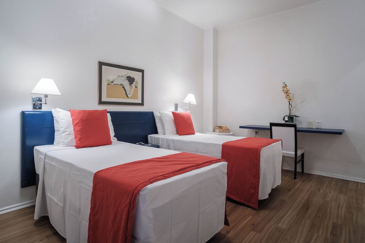 A hotel room with two beds, each with white bedding and red accents, a desk with a chair, and a framed picture on the wall.