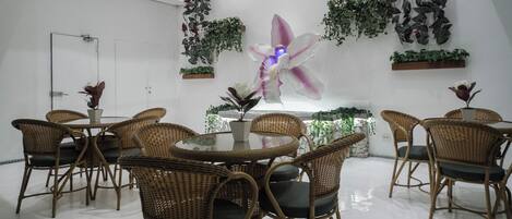 Un'area pranzo moderna con sedie in vimini e un tavolo in vetro, circondata da piante in vaso e un'ampia orchidea decorativa.