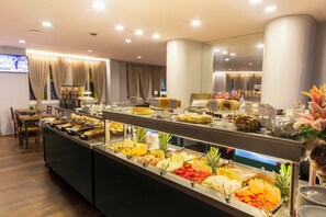 A buffet with various food items, a television screen, and a seating area with chairs and a table. - Hotel Normandie by Easy Plus (São Paulo)