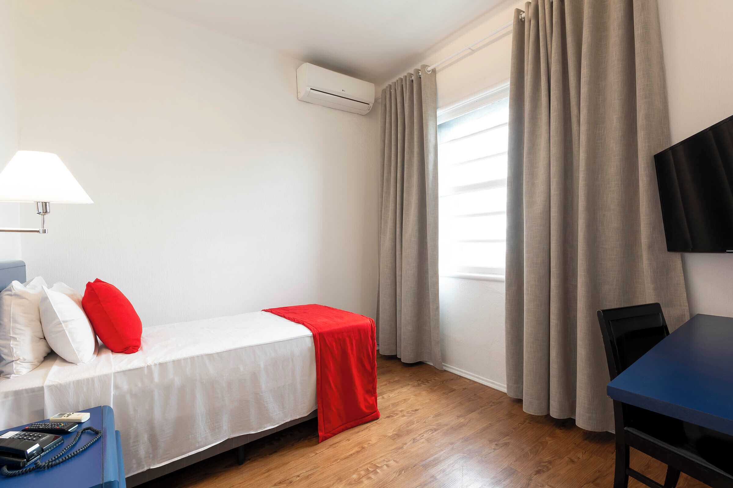 Habitación de hotel con cama, escritorio, silla, lámpara, aire acondicionado y televisión.