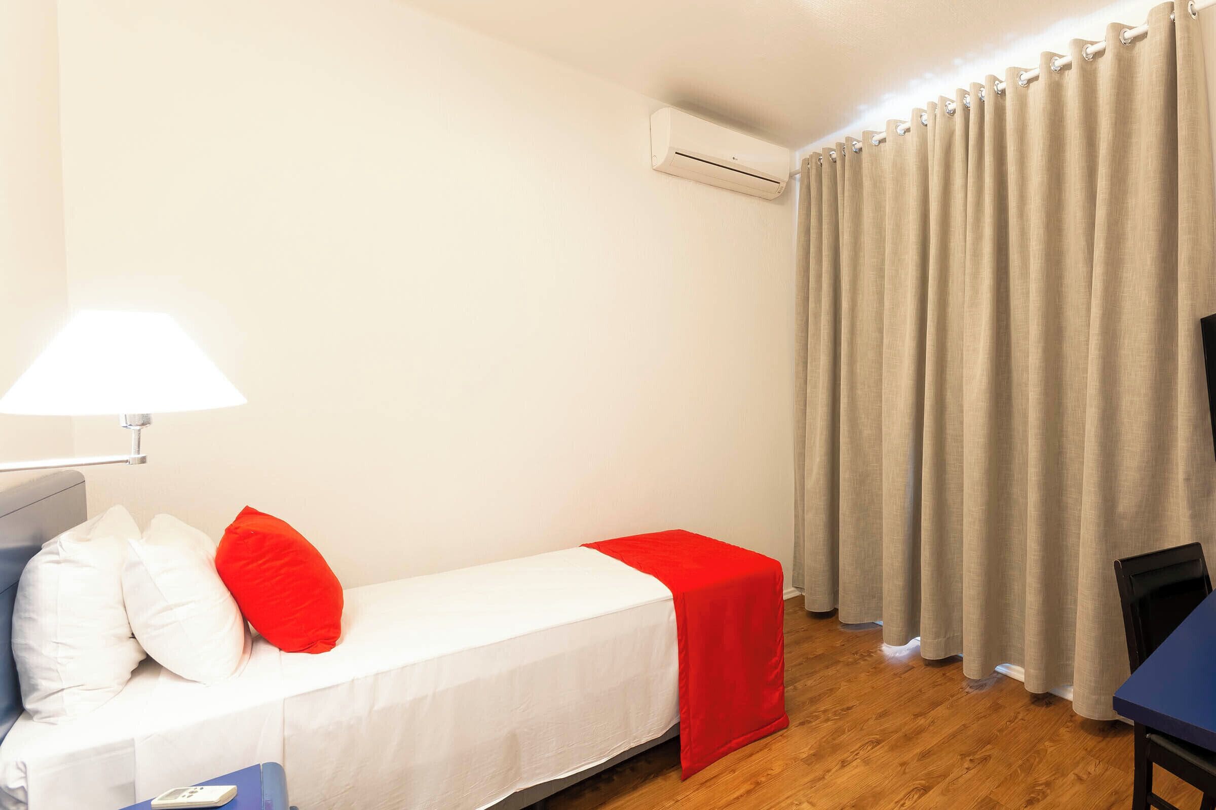 A small room with a single bed, a bedside table, a lamp, and a window with curtains.