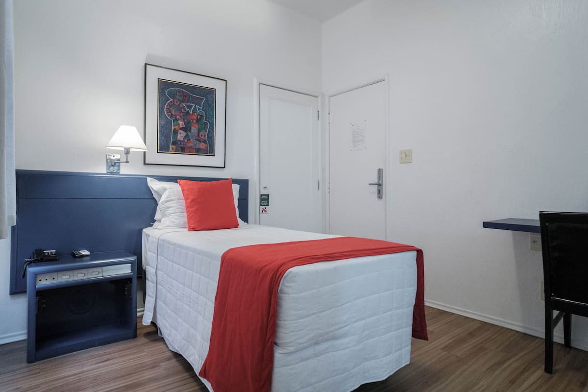 A hotel room with a bed, a desk, a chair, a nightstand, a lamp, and a framed picture on the wall.