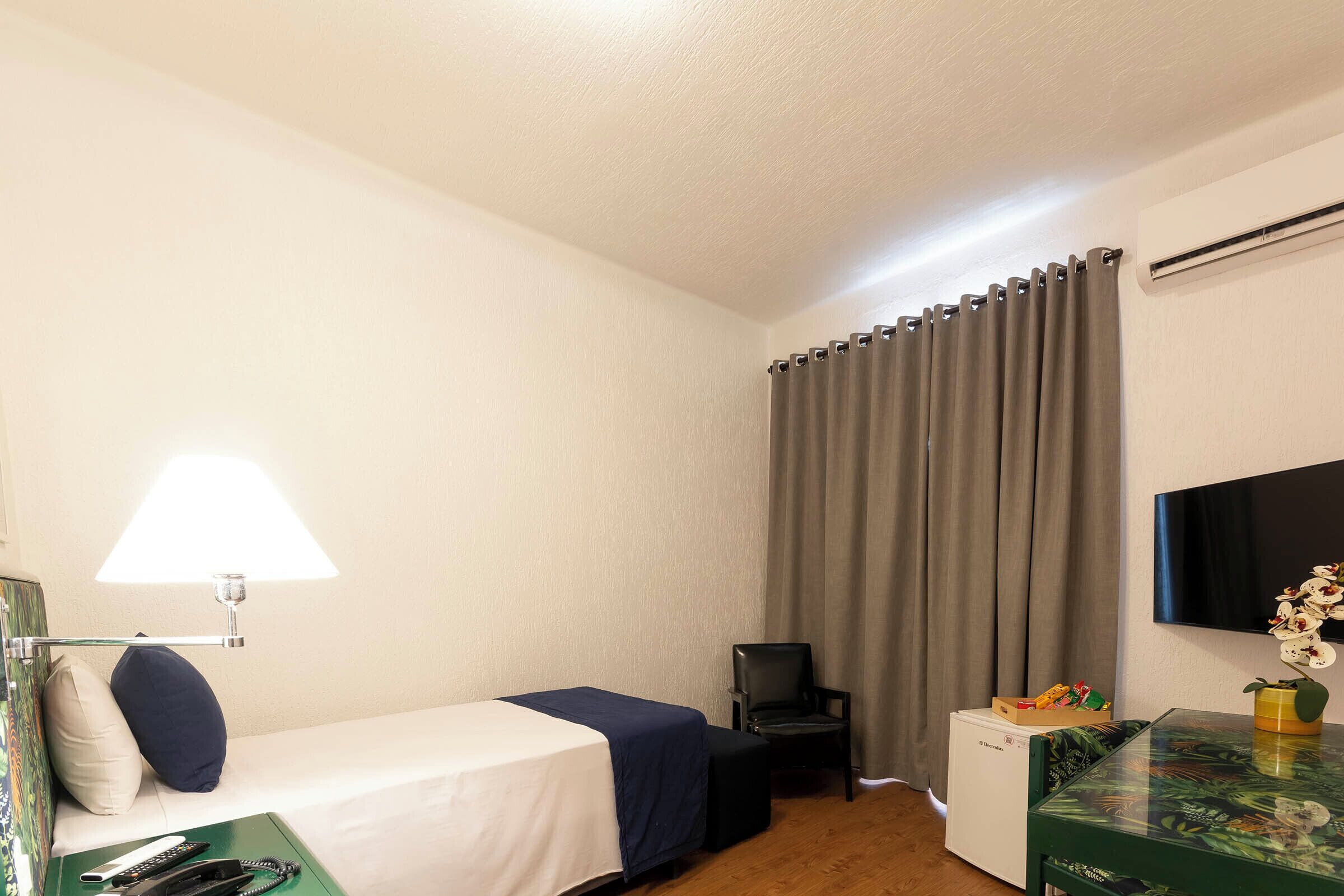 A hotel room with a bed, a desk with a lamp, a chair, a TV, and a small table with a vase.