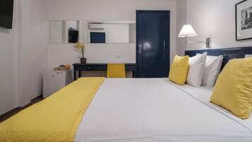 Habitación de hotel con cama, escritorio, silla, refrigerador pequeño, lámpara y puerta.