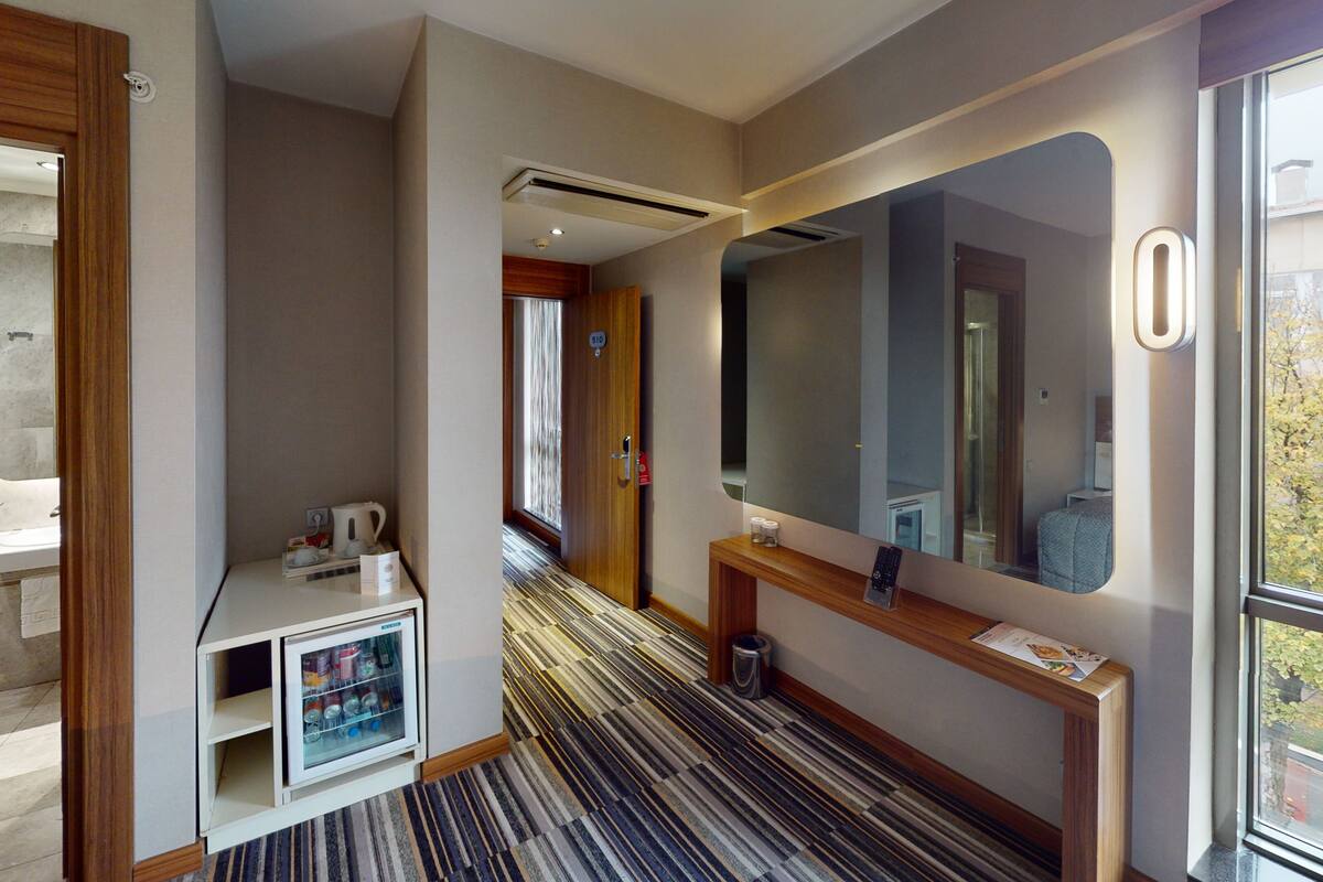 A hotel room with a bathroom, a minibar, a mirror, and a view of the outside.