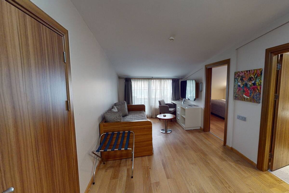 A modern hotel room with a wooden floor, a bed, a desk with a chair, a small table, and a shower area.