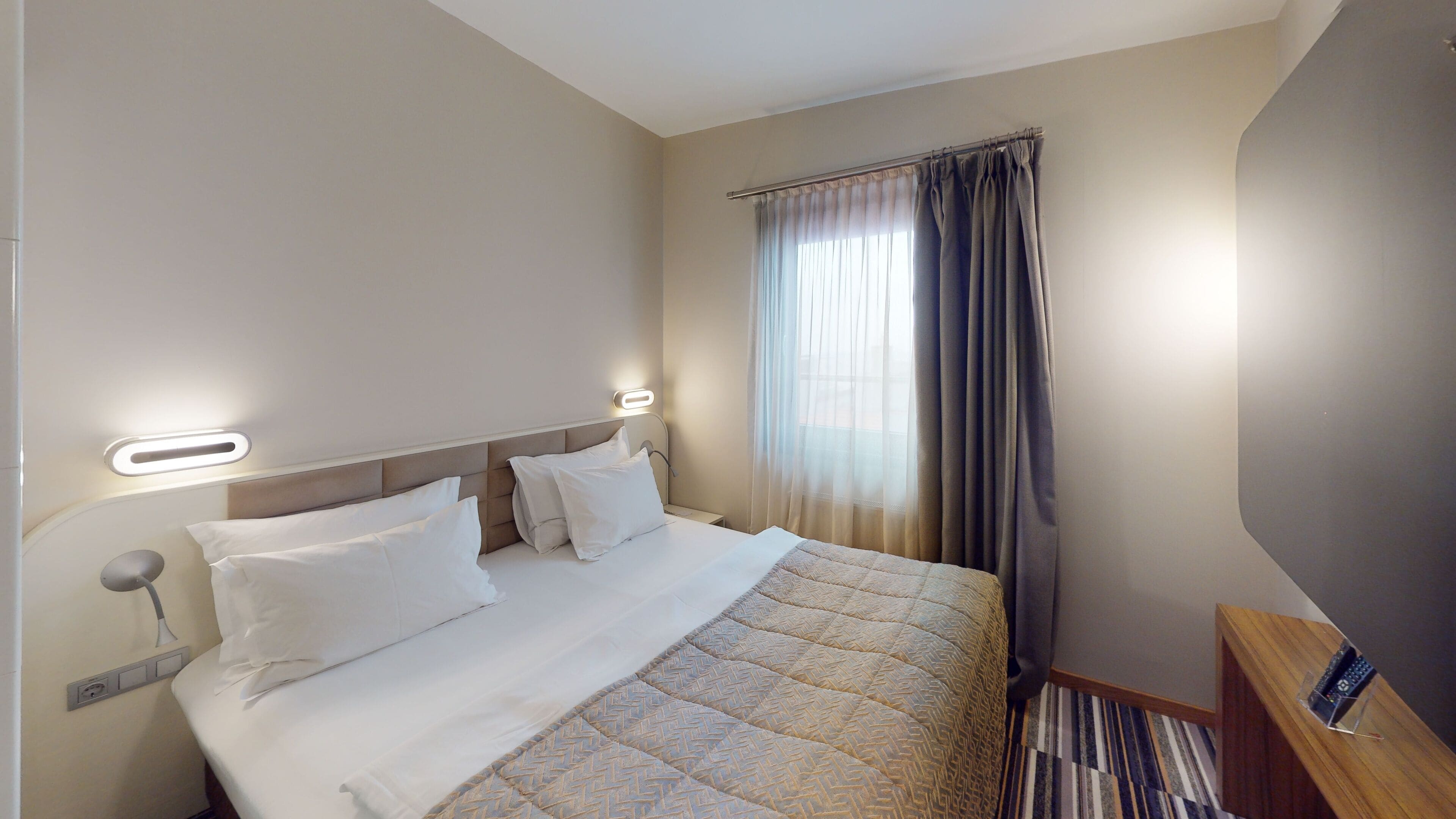 A hotel room with a bed, a bedside table, a nightstand, and a window with curtains.