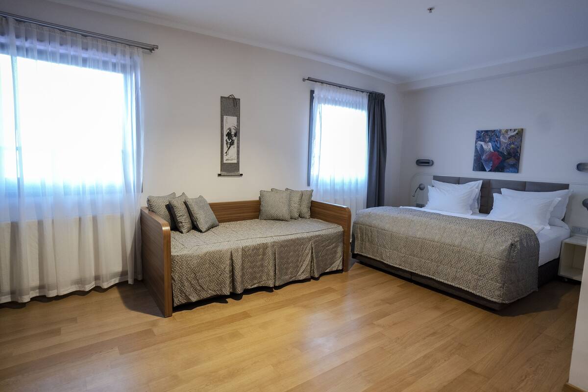A room with a sofa and a bed, both with patterned covers, wooden flooring, and curtains on the windows.