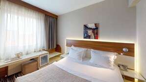 Quarto de hotel com cama, escrivaninha, cadeira, mesa de cabeceira, abajur e um quadro na parede.
