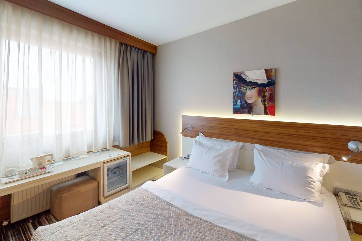 A hotel room with a bed, a desk, a chair, a bedside table, a lamp, and a painting on the wall.