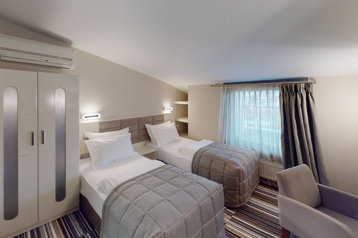 A hotel room with two beds, a wardrobe, a chair, a radiator, and a window with curtains.