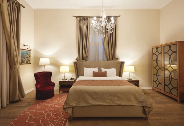 A hotel room with a large bed, two bedside tables with lamps, a red armchair, a wooden cabinet, and a chandelier.