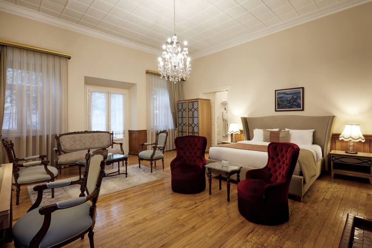 Ruime hotelkamer met een groot bed, een kroonluchter en klassiek meubilair, waaronder fauteuils en een bank.