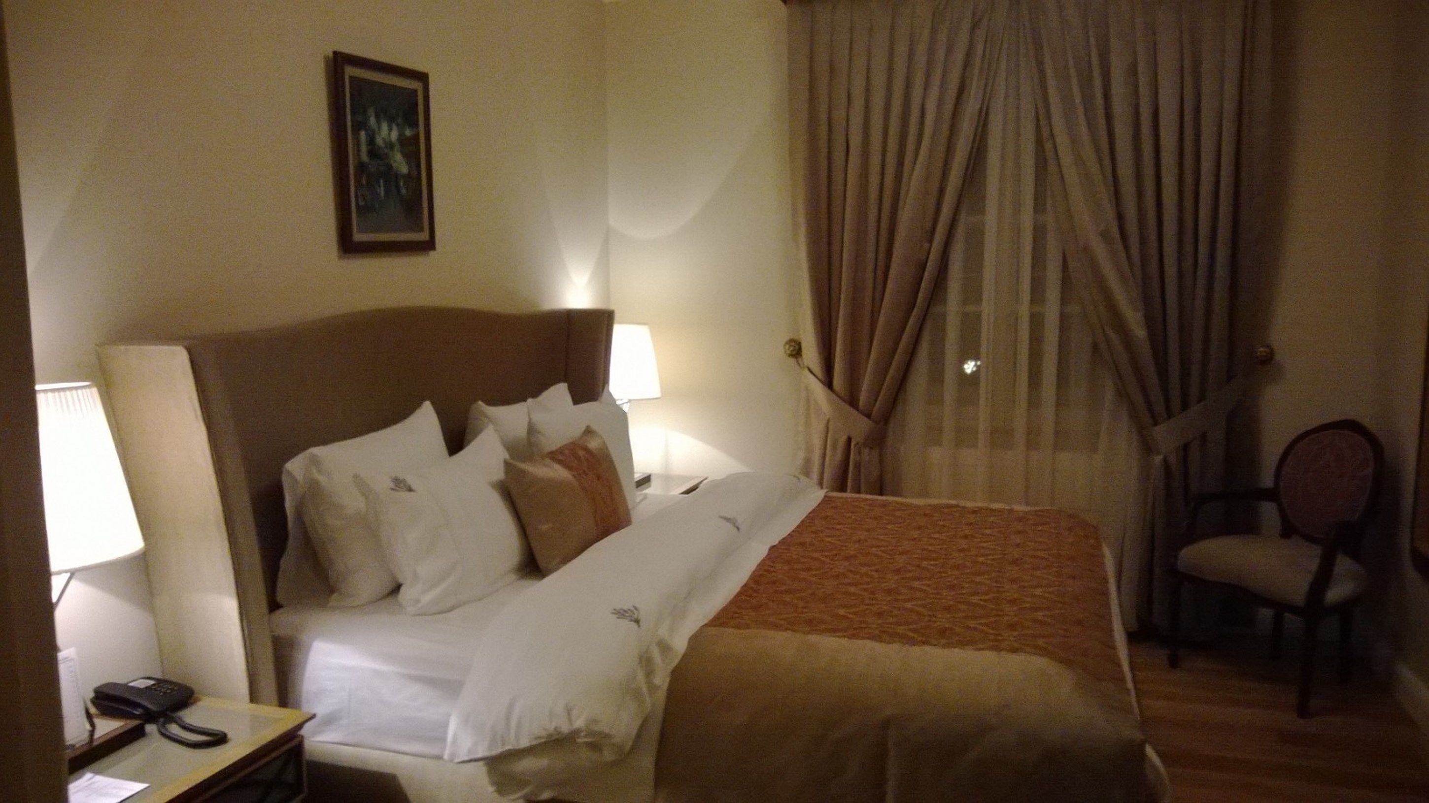 A hotel room with a bed, two bedside lamps, a chair, a telephone, and a painting on the wall.
