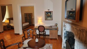Quarto de hotel com chão de madeira, uma mesa de jantar com cadeiras, lareira, cama, escrivaninha com candeeiro e um quadro na parede.