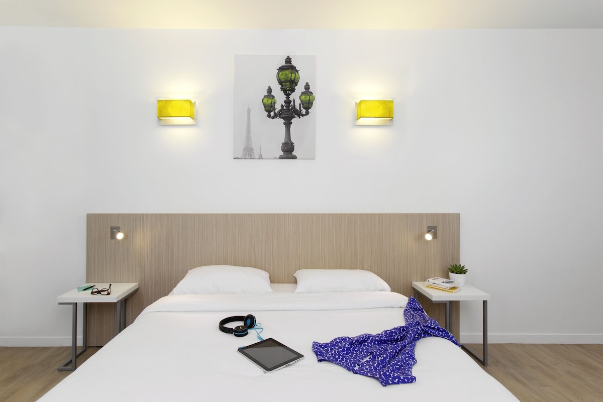 A hotel room with a bed, bedside tables, a lamp, a painting, and a wall-mounted light fixture.