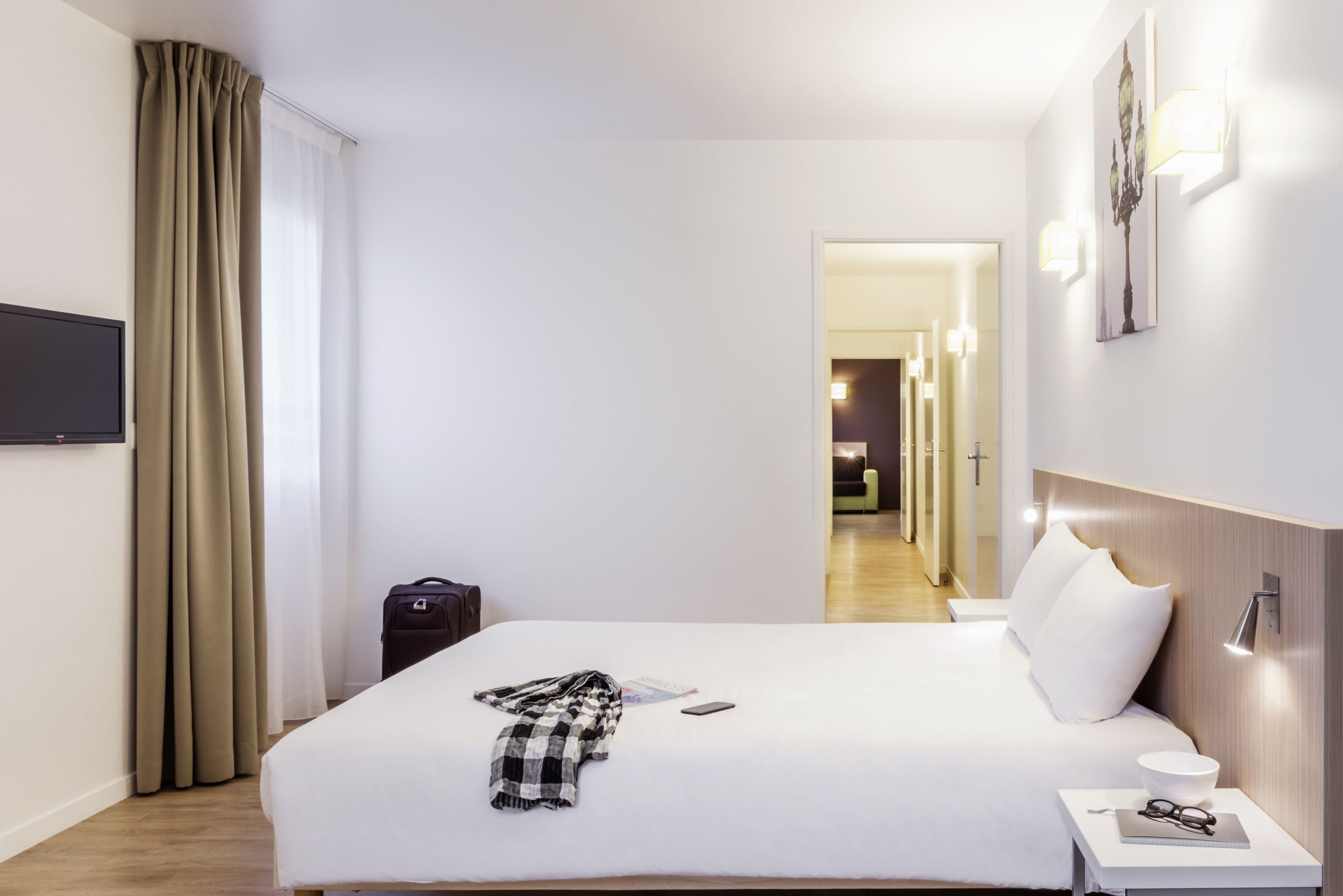 Una habitación de hotel con cama, mesita de noche, lámpara y vistas al pasillo.
