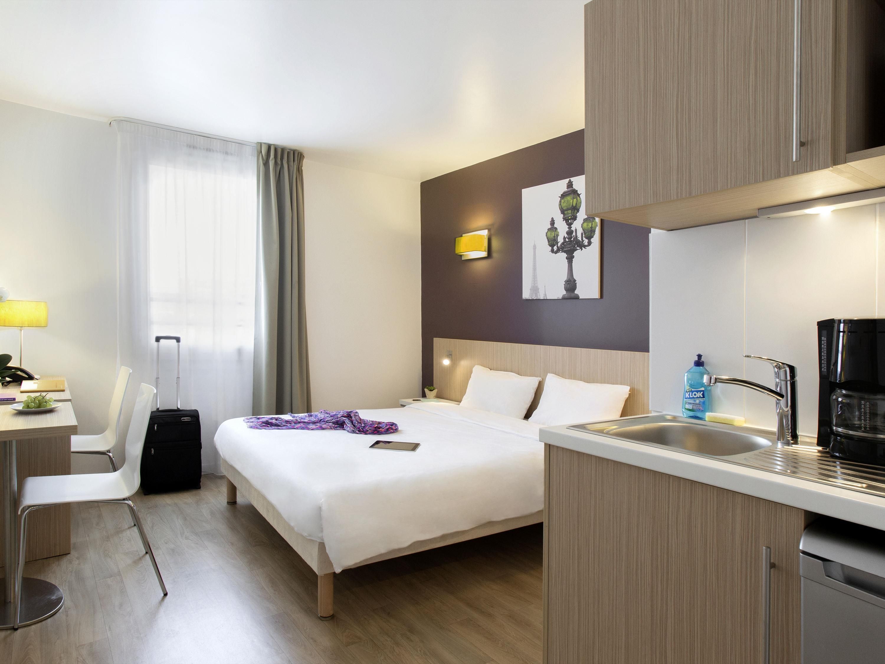 Une chambre d’hôtel comprenant un lit, une table de chevet, une lampe, une chaise, une valise et une kitchenette équipée d’un évier et d’une machine  à café.