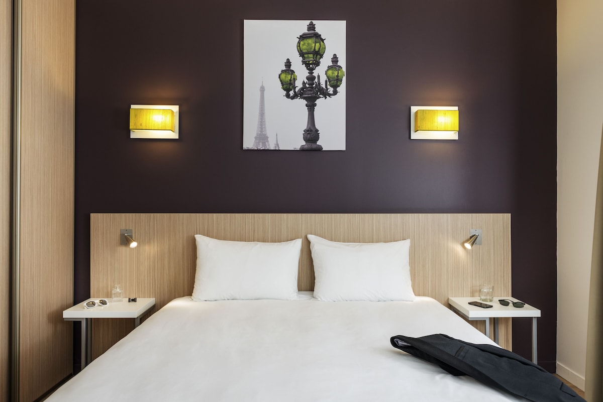 Une chambre d’hôtel avec un lit, deux tables de chevet, une petite table, une lampe fixée au mur et un tableau encadré représentant la Tour Eiffel.