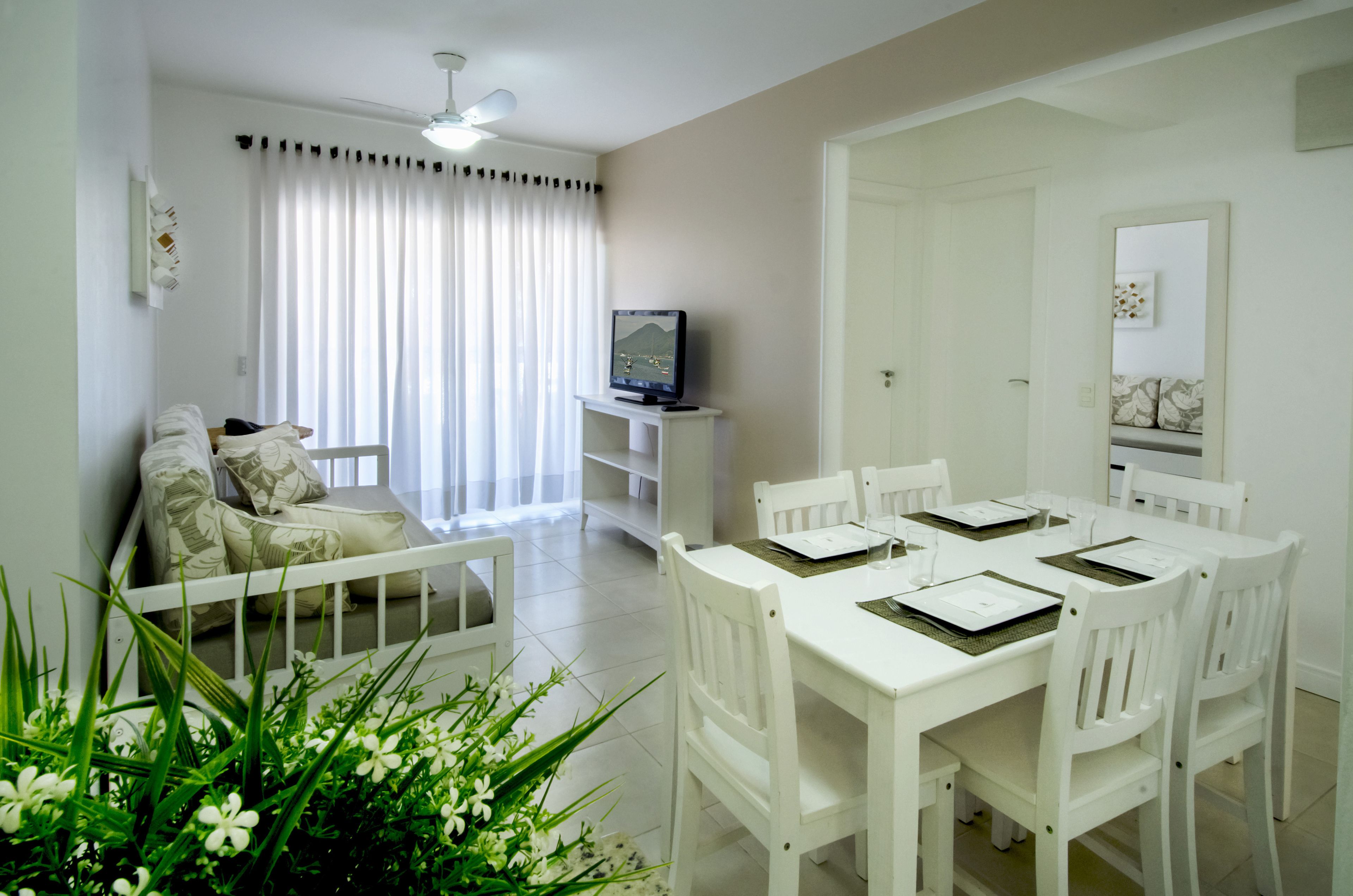 Een nette, witte woonkamer met een eettafel voor vier personen, een bank met gekleurde kussens, een televisie op een dressoir en een groot raam met gordijnen.
