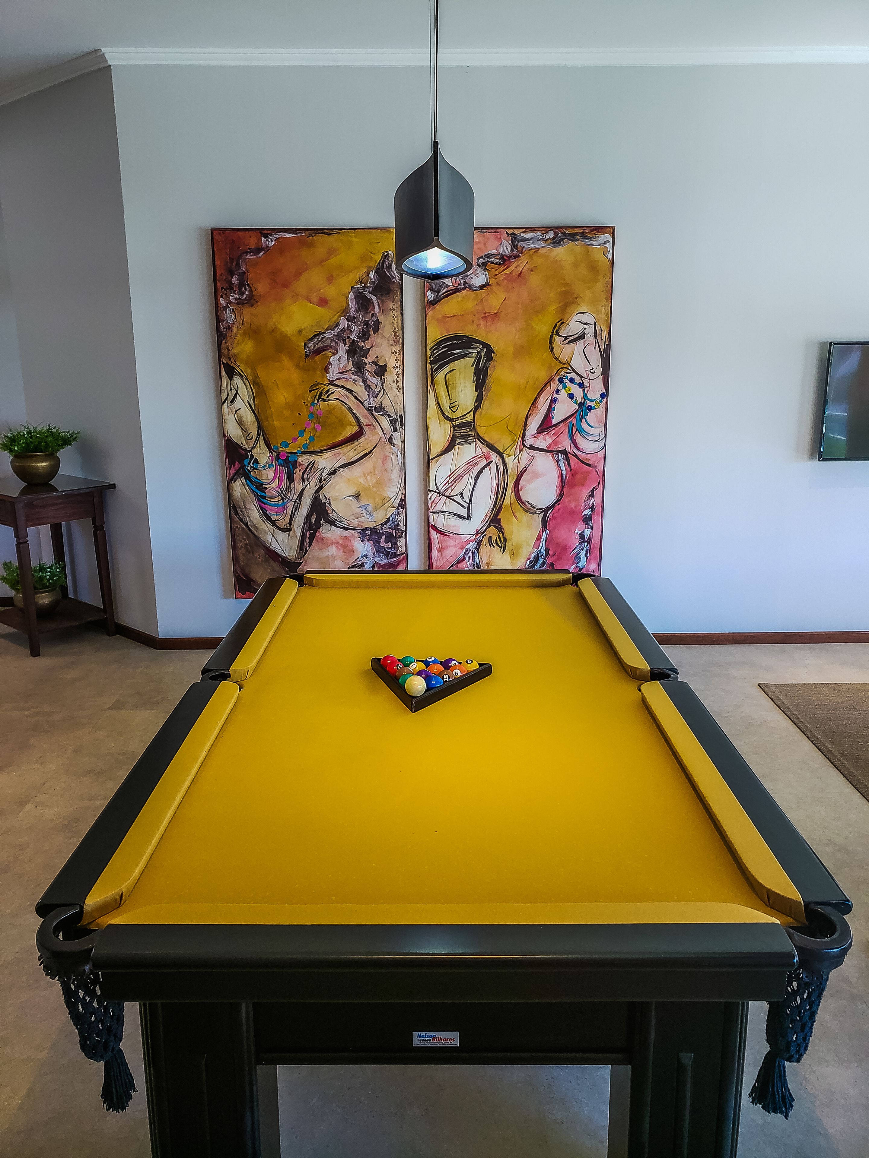A pool table with a yellow felt surface, a pool cue, and a decorative painting above it.